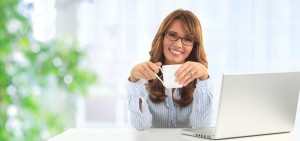 Attractive blond business woman working on her laptop and drinking coffee in her office (blue background)